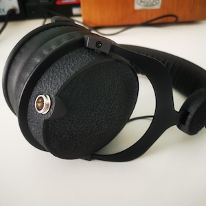 Könnte beinhalten: Schwarze Over-Ear-Kopfhörer mit einer texturierten, matten Oberfläche auf den runden Ohrmuscheln. Das Design umfasst einen metallischen Audio-Eingangsanschluss an der Seite, einen gepolsterten Kopfbügel und einen stabilen Rahmen.