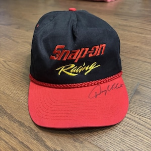 Può includere: Un cappellino da baseball nero con visiera rossa e un cordoncino intrecciato rosso alla base della corona. La parte anteriore presenta la scritta ricamata in rosso Snap-on e la scritta ricamata in giallo Racing. Una firma nera è sulla visiera.