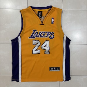 May include: A gold Los Angeles Lakers basketball jersey featuring the number 24 in silver with purple trim. The jersey has purple side panels with white vertical stripes, a V-neck collar, and an Adidas logo with an NBA emblem on the chest.