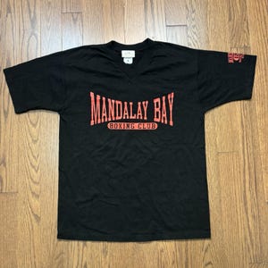 Puede incluir: Una camiseta negra de manga corta con cuello en V que presenta el texto rojo Mandalay Bay Boxing Club impreso en el pecho. La manga izquierda incluye un pequeño gráfico rojo con el número 12 y el texto Mandalay Bay Boxing Club.