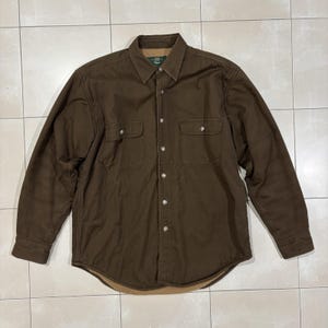 May include: A long-sleeved brown shirt featuring a pointed collar, a button-down front, and two chest pockets with buttoned flaps. The interior is lined with a tan fleece material, and the garment features metal buttons along the front placket.