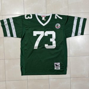 Könnte beinhalten: Ein dunkelgrünes Netz-Footballtrikot mit der weißen Nummer 73 auf der Vorderseite und den Schultern. Es hat einen weißen V-Ausschnitt, weiße Streifen an den Ärmeln, ein kreisförmiges Teamlogo auf der Brust und ein Etikett mit 1984 Joe Klecko.