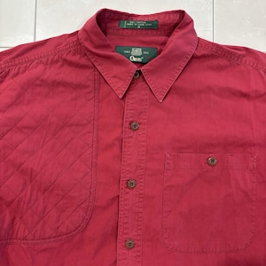 May include: A dark red button-down shirt featuring a quilted panel on the right side, a pointed collar, and a single chest pocket on the left. The interior label displays the text Orvis, since 1856, 100% cotton, and made in Hong Kong, size M.