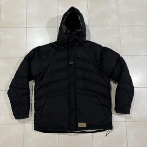 Puede incluir: Una chaqueta acolchada negra con capucha, con costuras horizontales, cierre de cremallera frontal y un pequeño logotipo de Oakley en el pecho. La chaqueta incluye dos bolsillos laterales y un parche rectangular cerca del dobladillo.