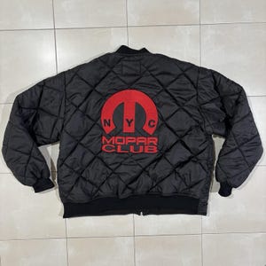 May include: A black quilted bomber jacket featuring a large red embroidered Mopar Club logo on the back. The design includes the letters NYC positioned within the red arch of the logo, with the words MOPAR CLUB printed in red underneath.