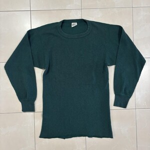 Puede incluir: Una camiseta de manga larga en color verde bosque oscuro con un tejido de textura de punto gofrado. La prenda presenta un cuello redondo clásico, puños acanalados en el extremo de cada manga y un dobladillo recto. Una pequeña etiqueta blanca es visible.