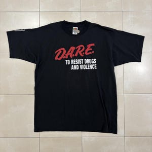 May include: A black short-sleeved t-shirt featuring the red text D.A.R.E. above the white text TO RESIST DRUGS AND VIOLENCE printed on the chest. The shirt has a crew neckline and a small white graphic detail on the edge of the right sleeve.