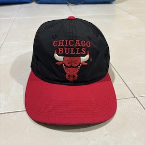 Op de afbeelding: Een zwarte honkbalpet met het Chicago Bulls-logo in rood en wit geborduurd op de voorkant. De pet heeft een effen rode klep met stikseldetails en een kleine rode knoop in het midden bovenop de kroon, met daarop de naam van het team.
