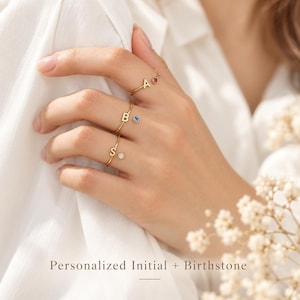 May include: Three thin gold rings, each featuring a single uppercase letter initial and a small round gemstone. The top ring has the letter A and a pink stone, the middle has B and a blue stone, and the bottom has S and a clear white stone.