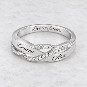Puede incluir: Un anillo de tono plateado con un diseño de infinito entrelazado y piedras claras incrustadas. La banda está grabada con los nombres Denton y Alex en la parte frontal, mientras que la superficie interior muestra la inscripción Love you forever.