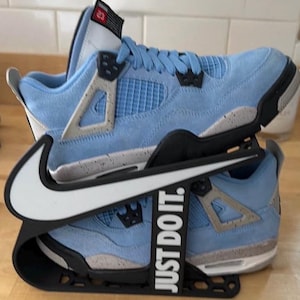 May include: A pair of light blue sneakers with grey speckled midsoles and black accents, displayed on a black plastic stand featuring a white Nike swoosh logo and the text "JUST DO IT." The sneakers have a mesh side panel and a tongue tag with "23."