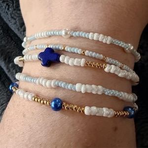 May include: A set of four beaded bracelets featuring a mix of white, light blue, and metallic gold seed beads. One bracelet includes a prominent dark blue cross-shaped bead, while another features two larger, round, glossy dark blue beads.