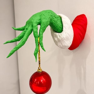 May include: A wall-mounted decoration featuring a textured green hand with long fingers emerging from a red sleeve with a white fur-like cuff. The hand is holding a shiny, spherical red Christmas ornament by a thin gold string.