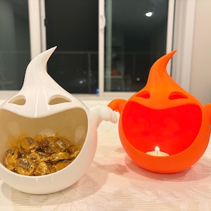 May include: Two ghost-shaped bowls, one white and one orange, featuring wide open mouths and small arms. The white bowl is filled with gold-wrapped sweets, while the orange bowl contains a small, lit white tea light candle inside its hollow centre.