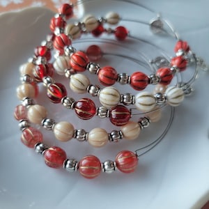 May include: A collection of beaded bracelets. The bracelets feature red, cream, and clear beads with silver accents. The beads are strung on a thin wire, creating a delicate and elegant look. The bracelets are arranged in a circular pattern.