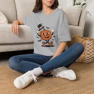 May include: A heather gray short-sleeve t-shirt featuring a graphic of an orange pumpkin character wearing a black top hat and brown boots. The pumpkin has arms and legs, and is surrounded by small black bats and a spider illustration.