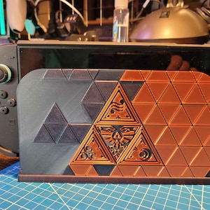 Op de afbeelding: Een zwart en koperkleurig 3D-geprint dockingstation voor een gameconsole met een geometrisch driehoekspatroon. Het midden toont een grote driehoek met een vogelembleem, geflankeerd door twee kleinere driehoeken met cirkelvormige symbolen.