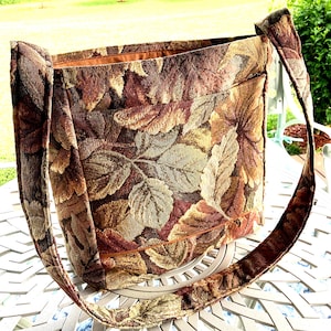 May include: A rectangular shoulder bag crafted from a woven tapestry fabric featuring a dense pattern of autumn-toned leaves in shades of brown, gold, rust, and tan. The bag includes a wide shoulder strap and a horizontal exterior pocket.