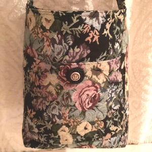 May include: A rectangular crossbody bag made of woven tapestry fabric featuring a dark background with a multi-colored floral pattern of roses and leaves. The bag includes a front pocket secured by a single round black button with a gold center.