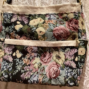 May include: A rectangular shoulder bag featuring a woven tapestry fabric with a multi-coloured floral pattern on a black background. The bag includes a cream-coloured fabric trim along the top and a horizontal cream-coloured pocket across the front.