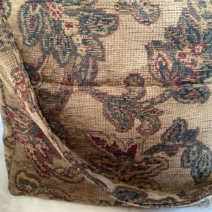 May include: A shoulder bag crafted from a textured, woven fabric featuring a multi-coloured floral pattern in shades of tan, dark green, and deep red. The bag includes a wide shoulder strap and a horizontal zippered pocket on the front exterior.
