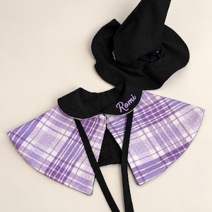 May include: A black pointed witch hat and a purple and white plaid cape with a black collar. The collar features the name Romi embroidered in purple script. The cape includes two long black fabric ties for fastening at the front.