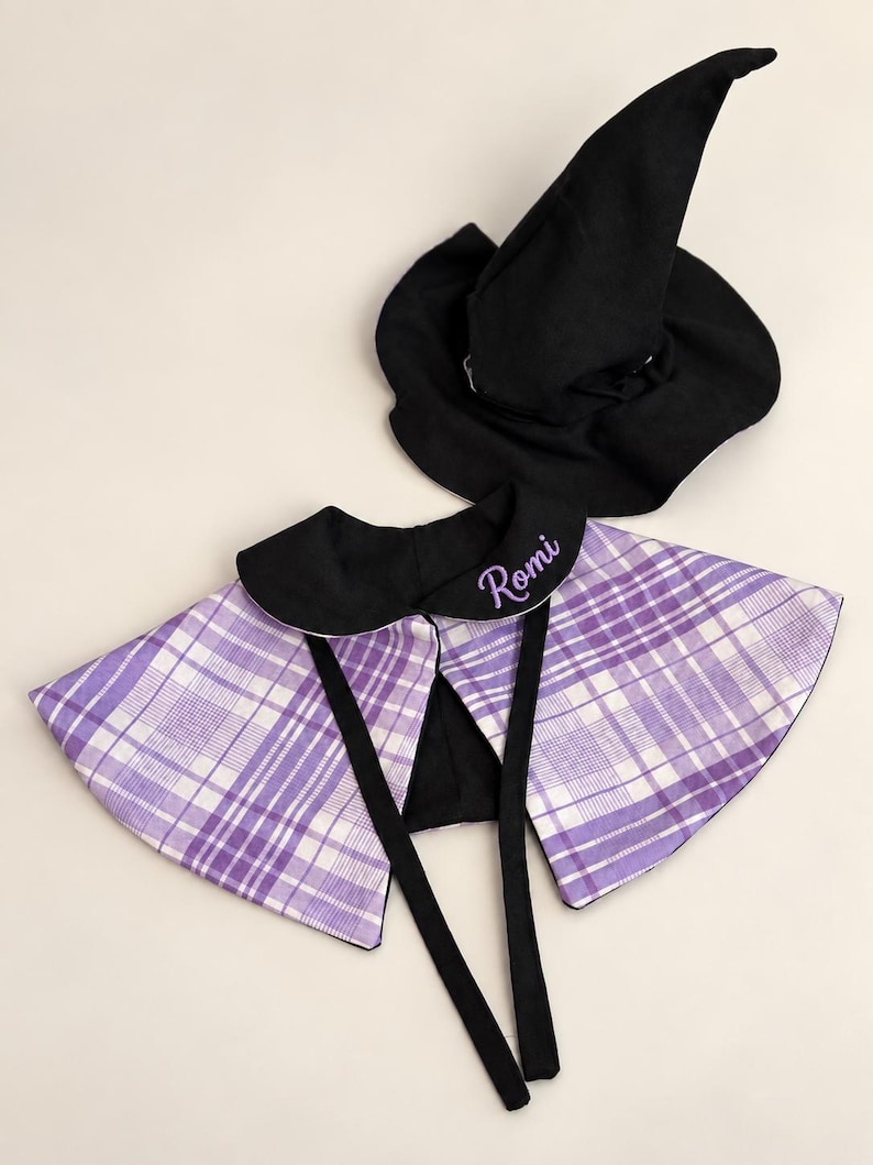 May include: A black pointed witch hat and a purple and white plaid cape with a black collar. The collar features the name Romi embroidered in purple script. The cape includes two long black fabric ties for fastening at the front.