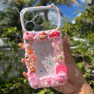 Peut inclure: Une coque de t&eacute;l&eacute;phone transparente d&eacute;cor&eacute;e d'une texture rose fa&ccedil;on cr&egrave;me fouett&eacute;e, orn&eacute;e de diverses breloques miniatures dont un ours, un champignon, un cupcake et un c&oelig;ur. Le centre est rempli de confettis en forme d'&eacute;toiles et de paillettes.
