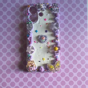 Peut inclure: Une coque de t&eacute;l&eacute;phone rectangulaire avec une surface blanche textur&eacute;e fa&ccedil;on cr&egrave;me, bord&eacute;e de gla&ccedil;age violet. Elle est orn&eacute;e de breloques vari&eacute;es, dont un ours, un papillon, des &eacute;toiles, des fleurs, un c&oelig;ur et des vermicelles color&eacute;s.