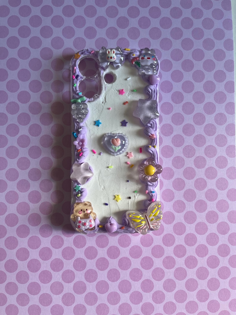 Peut inclure: Une coque de t&eacute;l&eacute;phone rectangulaire avec une surface blanche textur&eacute;e fa&ccedil;on cr&egrave;me, bord&eacute;e de gla&ccedil;age violet. Elle est orn&eacute;e de breloques vari&eacute;es, dont un ours, un papillon, des &eacute;toiles, des fleurs, un c&oelig;ur et des vermicelles color&eacute;s.