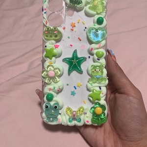 Peut inclure: Une coque de t&eacute;l&eacute;phone transparente d&eacute;cor&eacute;e de bordures en cr&egrave;me fouett&eacute;e vert clair. Le design pr&eacute;sente divers charms en r&eacute;sine, notamment une &eacute;toile de mer verte, une grenouille, un ours, un papillon, des fleurs et des vermicelles color&eacute;s.