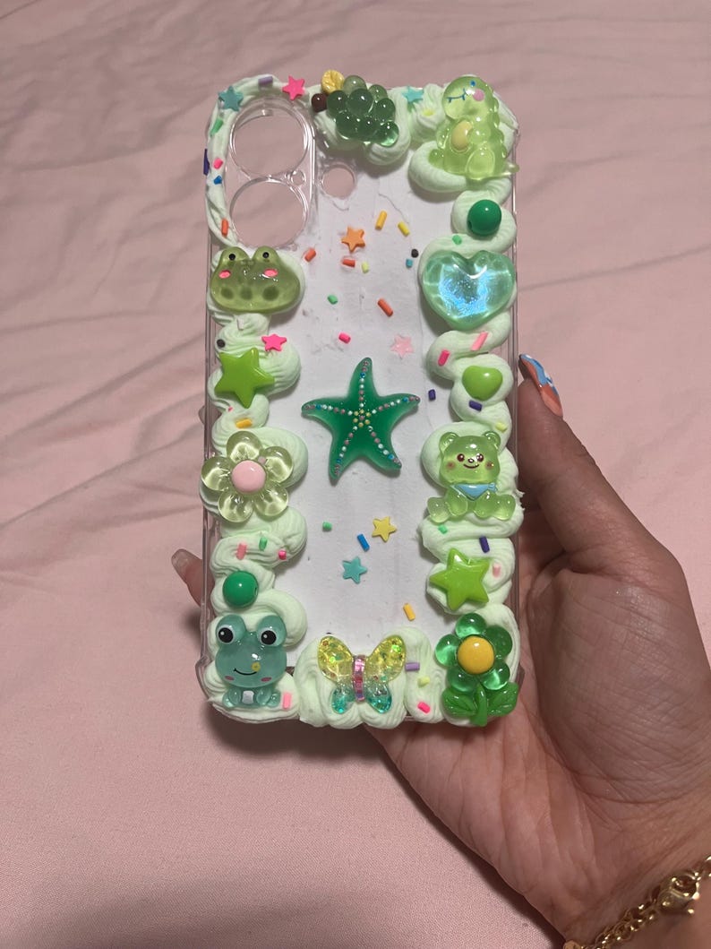 Peut inclure: Une coque de t&eacute;l&eacute;phone transparente d&eacute;cor&eacute;e de bordures en cr&egrave;me fouett&eacute;e vert clair. Le design pr&eacute;sente divers charms en r&eacute;sine, notamment une &eacute;toile de mer verte, une grenouille, un ours, un papillon, des fleurs et des vermicelles color&eacute;s.