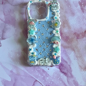 Peut inclure: Une coque de t&eacute;l&eacute;phone bleu clair avec une surface textur&eacute;e fa&ccedil;on matelass&eacute;e incrust&eacute;e de petits strass transparents. Les bords sont orn&eacute;s d'un passepoil blanc rappelant la cr&egrave;me fouett&eacute;e et de diverses breloques miniatures.