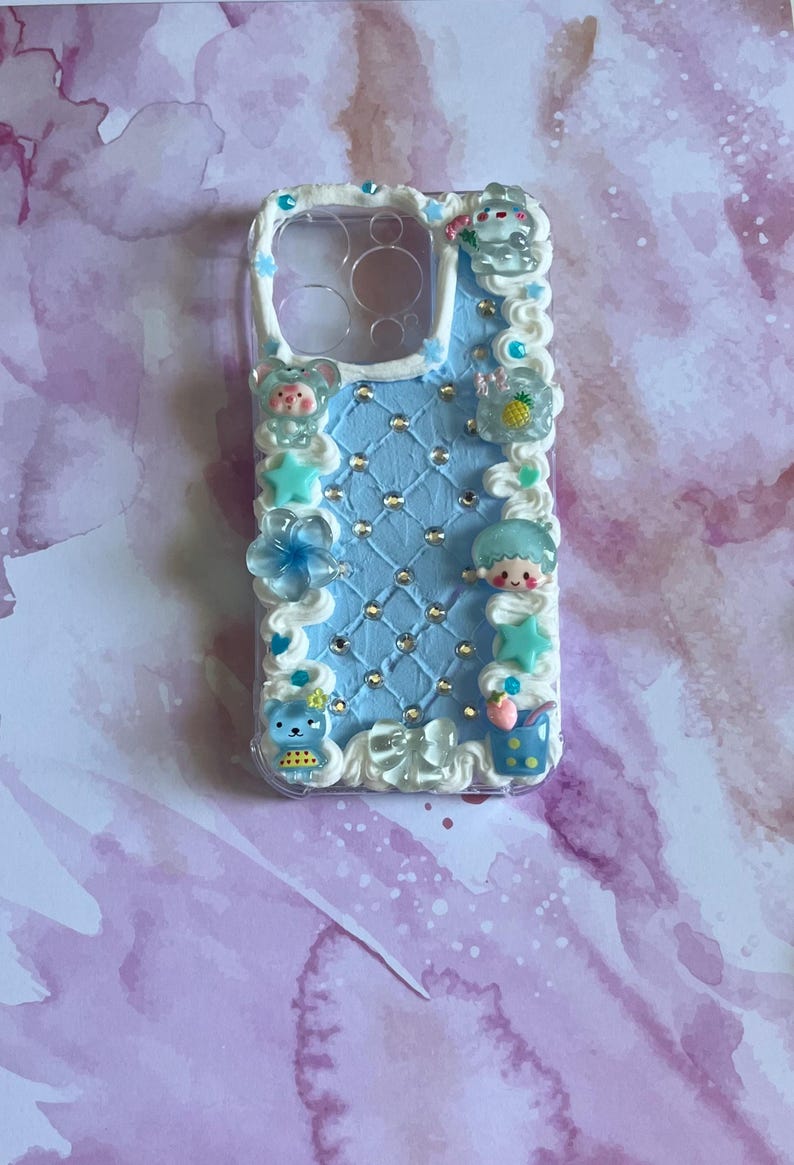 Peut inclure: Une coque de t&eacute;l&eacute;phone bleu clair avec une surface textur&eacute;e fa&ccedil;on matelass&eacute;e incrust&eacute;e de petits strass transparents. Les bords sont orn&eacute;s d'un passepoil blanc rappelant la cr&egrave;me fouett&eacute;e et de diverses breloques miniatures.