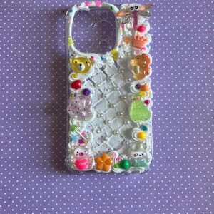 Peut inclure: Une coque de t&eacute;l&eacute;phone transparente d&eacute;cor&eacute;e d'une texture fa&ccedil;on cr&egrave;me fouett&eacute;e blanche, de perles blanches et de divers charms color&eacute;s, dont une libellule, un ours, une couronne, une bouteille verte, une fleur et un petit c&oelig;ur avec le mot LOVE.