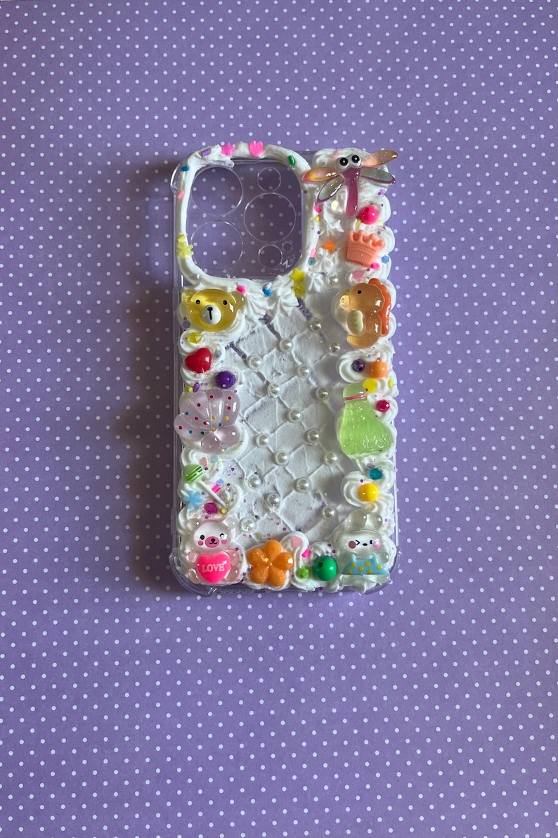 Peut inclure: Une coque de t&eacute;l&eacute;phone transparente d&eacute;cor&eacute;e d'une texture fa&ccedil;on cr&egrave;me fouett&eacute;e blanche, de perles blanches et de divers charms color&eacute;s, dont une libellule, un ours, une couronne, une bouteille verte, une fleur et un petit c&oelig;ur avec le mot LOVE.