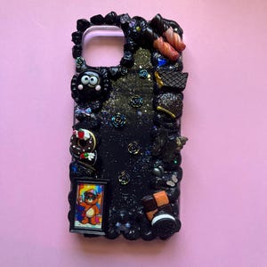 Peut inclure: Une coque de t&eacute;l&eacute;phone noire orn&eacute;e d'une finition paillet&eacute;e sur le th&egrave;me de la galaxie et de diverses breloques miniatures, dont une petite illustration d'ours encadr&eacute;e, un beignet, un biscuit et plusieurs roses sombres sur les bords.