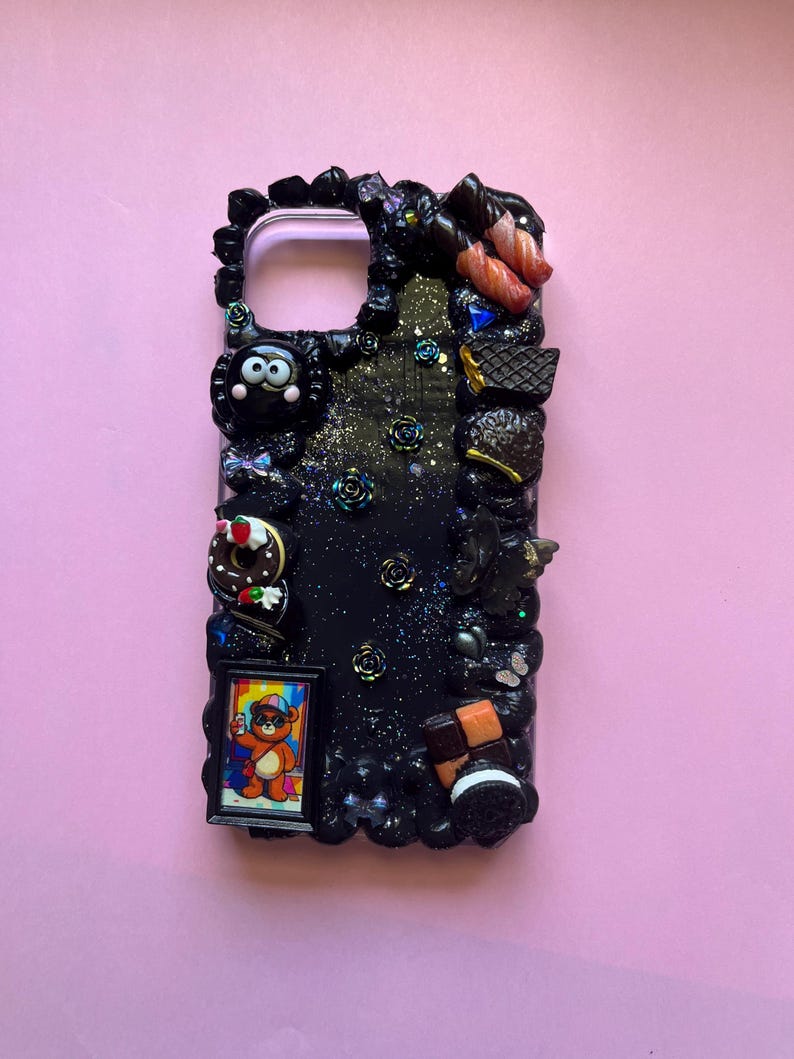 Peut inclure: Une coque de t&eacute;l&eacute;phone noire orn&eacute;e d'une finition paillet&eacute;e sur le th&egrave;me de la galaxie et de diverses breloques miniatures, dont une petite illustration d'ours encadr&eacute;e, un beignet, un biscuit et plusieurs roses sombres sur les bords.