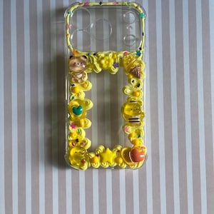 Peut inclure: Une coque de t&eacute;l&eacute;phone transparente d&eacute;cor&eacute;e d'une texture jaune semblable &agrave; de la cr&egrave;me fouett&eacute;e, orn&eacute;e de breloques en r&eacute;sine dont un ours, une abeille, un cornet de glace, une &eacute;toile et un c&oelig;ur, avec des paillettes color&eacute;es sur les bords.