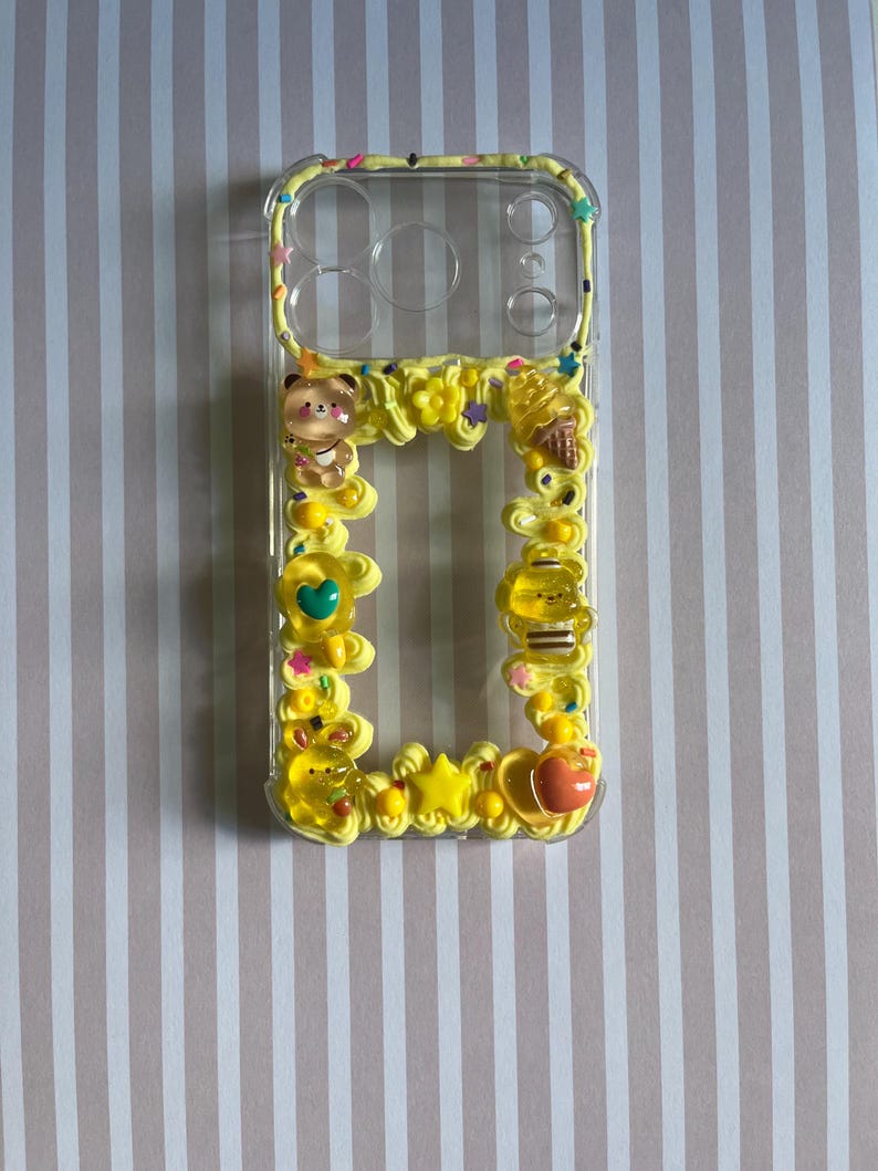 Peut inclure: Une coque de t&eacute;l&eacute;phone transparente d&eacute;cor&eacute;e d'une texture jaune semblable &agrave; de la cr&egrave;me fouett&eacute;e, orn&eacute;e de breloques en r&eacute;sine dont un ours, une abeille, un cornet de glace, une &eacute;toile et un c&oelig;ur, avec des paillettes color&eacute;es sur les bords.