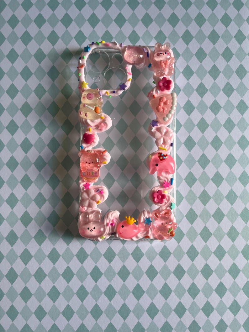 Peut inclure: Une coque de t&eacute;l&eacute;phone transparente d&eacute;cor&eacute;e d'une texture rose fa&ccedil;on cr&egrave;me fouett&eacute;e, orn&eacute;e de divers charms en r&eacute;sine color&eacute;s, notamment des ours translucides, des fleurs, des &eacute;toiles et des petites paillettes autour du contour.