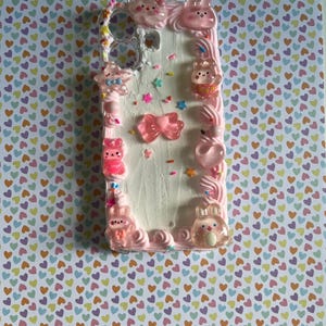 Peut inclure: Une coque de t&eacute;l&eacute;phone rectangulaire avec une surface blanche textur&eacute;e rappelant le gla&ccedil;age, d&eacute;cor&eacute;e de chantilly rose sur les bords, de vermicelles color&eacute;s et de divers charms roses translucides incluant des lapins, des &eacute;toiles et un n&oelig;ud central.