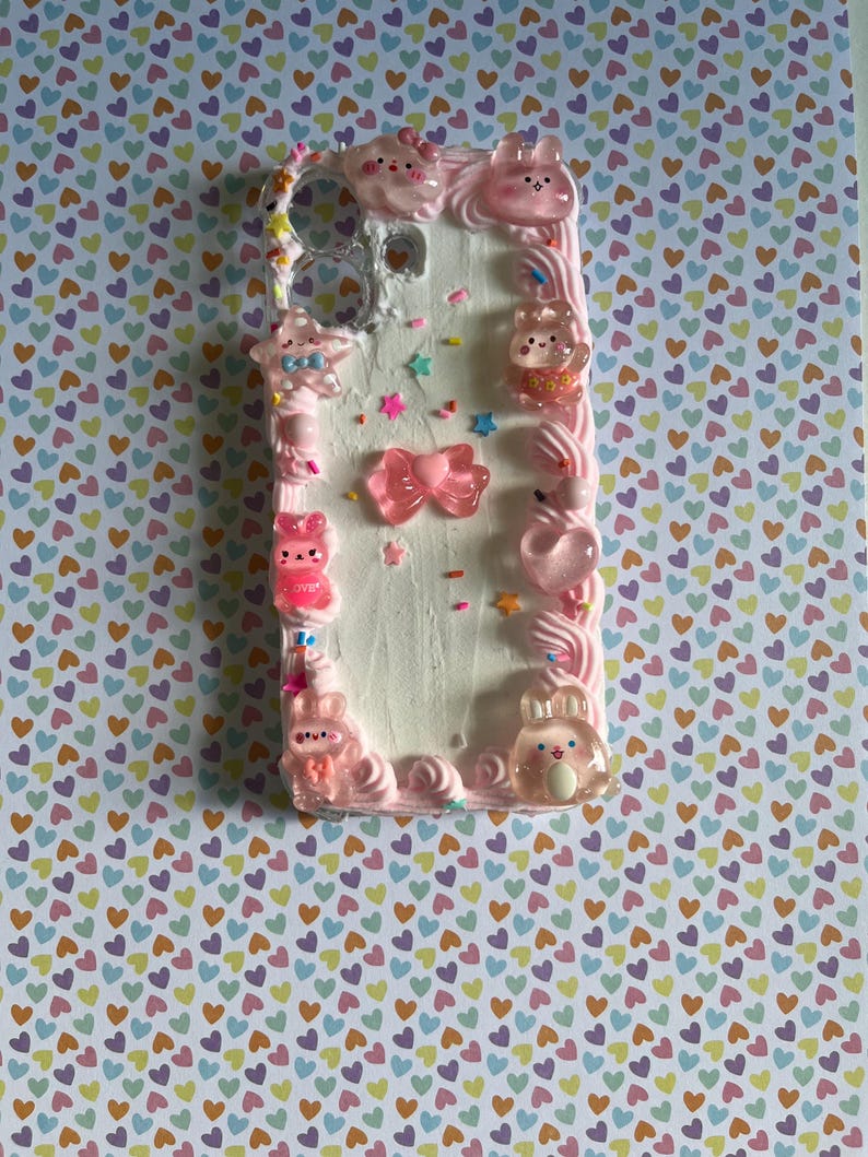 Peut inclure: Une coque de t&eacute;l&eacute;phone rectangulaire avec une surface blanche textur&eacute;e rappelant le gla&ccedil;age, d&eacute;cor&eacute;e de chantilly rose sur les bords, de vermicelles color&eacute;s et de divers charms roses translucides incluant des lapins, des &eacute;toiles et un n&oelig;ud central.