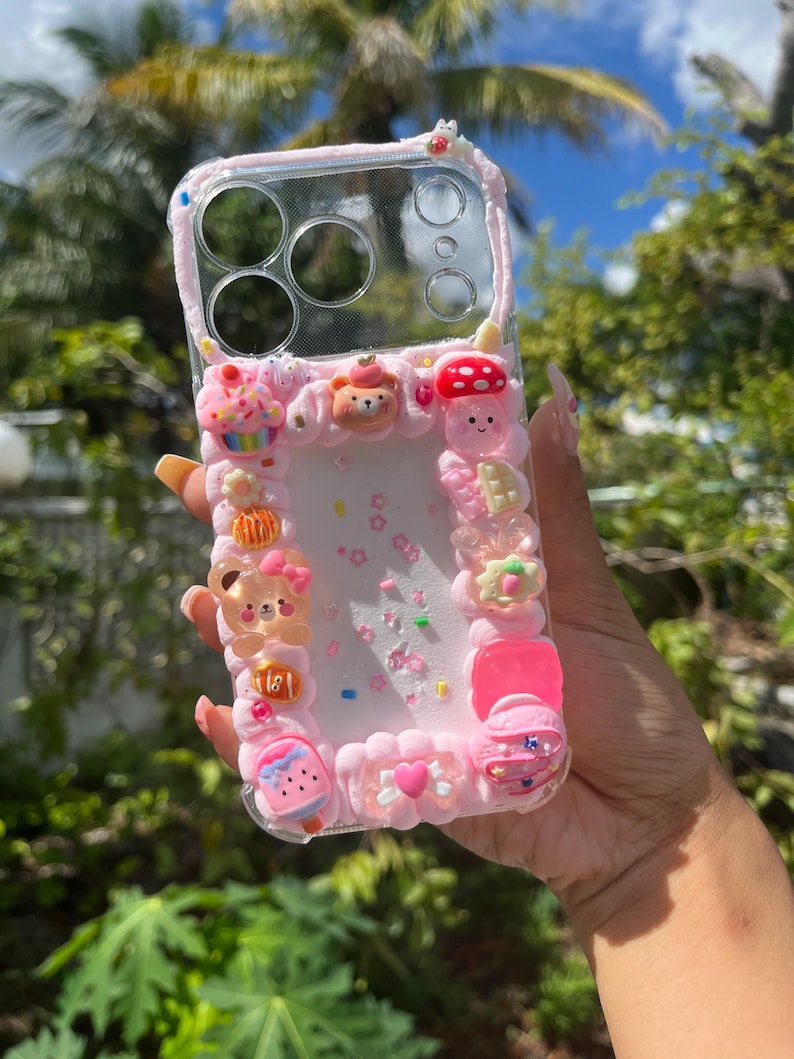Peut inclure: Une coque de t&eacute;l&eacute;phone transparente d&eacute;cor&eacute;e d'une texture rose fa&ccedil;on cr&egrave;me fouett&eacute;e, orn&eacute;e de diverses breloques miniatures dont un ours, un champignon, un cupcake et un c&oelig;ur. Le centre est rempli de confettis en forme d'&eacute;toiles et de paillettes.