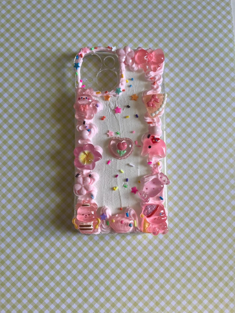 Peut inclure: Une coque de t&eacute;l&eacute;phone transparente d&eacute;cor&eacute;e d'une texture ressemblant &agrave; de la cr&egrave;me fouett&eacute;e blanche, de vermicelles color&eacute;s et de divers charms en r&eacute;sine rose, dont un n&oelig;ud, un c&oelig;ur avec une fleur, un ours et d'autres petites figurines.