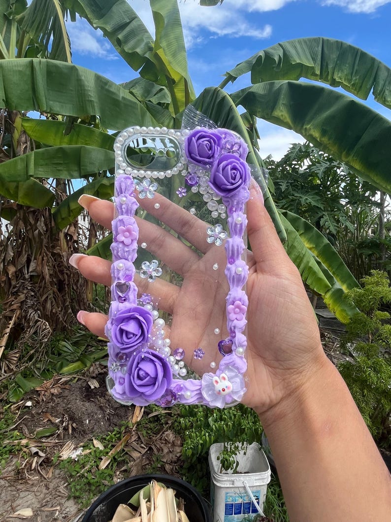 Peut inclure: Une coque de t&eacute;l&eacute;phone transparente orn&eacute;e de fausses roses violettes, de perles blanches et d'une texture cr&egrave;me violette. Le design comprend de petits cristaux en forme de fleurs, des c&oelig;urs et une petite figurine de lapin blanc en bas.