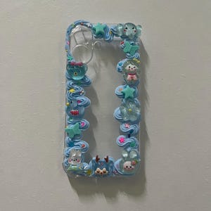 Peut inclure: Une coque de t&eacute;l&eacute;phone transparente d&eacute;cor&eacute;e de bordures en cr&egrave;me chantilly bleu clair. Le cadre pr&eacute;sente divers petits charms en r&eacute;sine, notamment un ours bleu, un papillon vert, une &eacute;toile, une fleur et plusieurs petites figurines d'animaux.