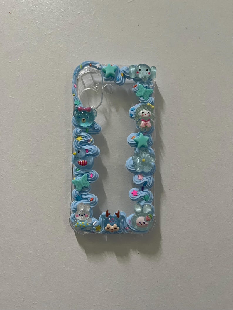 Peut inclure: Une coque de t&eacute;l&eacute;phone transparente d&eacute;cor&eacute;e de bordures en cr&egrave;me chantilly bleu clair. Le cadre pr&eacute;sente divers petits charms en r&eacute;sine, notamment un ours bleu, un papillon vert, une &eacute;toile, une fleur et plusieurs petites figurines d'animaux.
