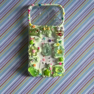 Peut inclure: Une coque de t&eacute;l&eacute;phone transparente d&eacute;cor&eacute;e d'une texture fa&ccedil;on cr&egrave;me fouett&eacute;e vert clair, orn&eacute;e de divers charms en r&eacute;sine color&eacute;s dont une grenouille, un ours, des fleurs, des &eacute;toiles et un petit n&oelig;ud, avec des paillettes multicolores.