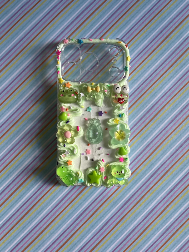 Peut inclure: Une coque de t&eacute;l&eacute;phone transparente d&eacute;cor&eacute;e d'une texture fa&ccedil;on cr&egrave;me fouett&eacute;e vert clair, orn&eacute;e de divers charms en r&eacute;sine color&eacute;s dont une grenouille, un ours, des fleurs, des &eacute;toiles et un petit n&oelig;ud, avec des paillettes multicolores.